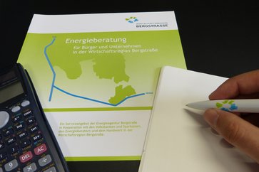 Symbolbild: Taschenrechner, Informationsbroschüre über Energieberatung sowie ein Noitzblock und eine Hand, welche einen Stift hält